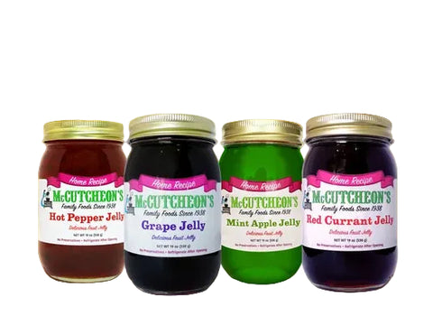 Four jars of McCutcheon's Family Farms Delicious Fruit Jelly arranged on a wooden surface with a blurred outdoor garden background. The jars include Hot Pepper Jelly, Grape Jelly, Mint Apple Jelly, and Red Currant Jelly.