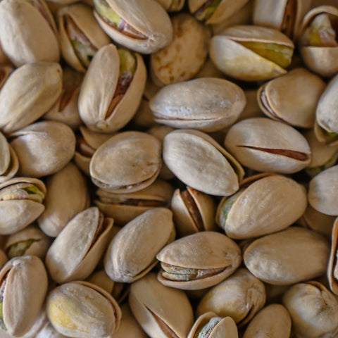 A close-up pile of shelled pistachios filling the frame, showing pale beige shells with some green pistachio visible in the splits.