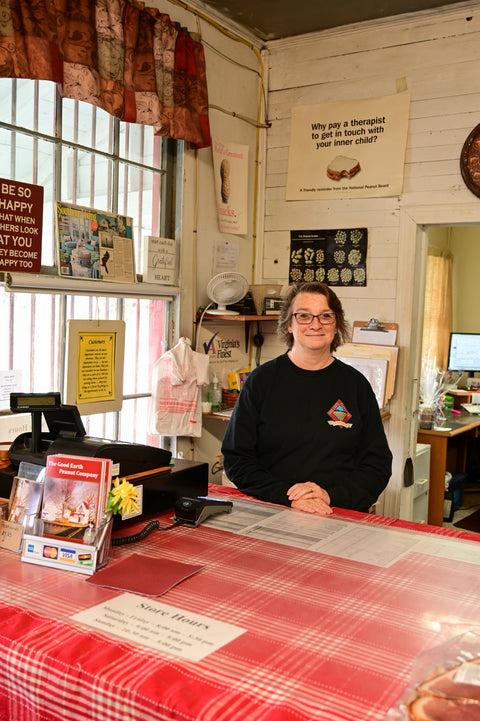 Person standing behind a counter in a small store with various items and signs on the walls.