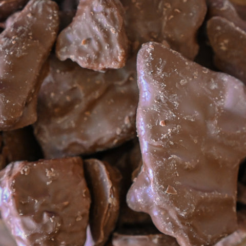 A close-up pile of chocolate-covered peanut brittle with a glossy brown coating and small textured inclusions.