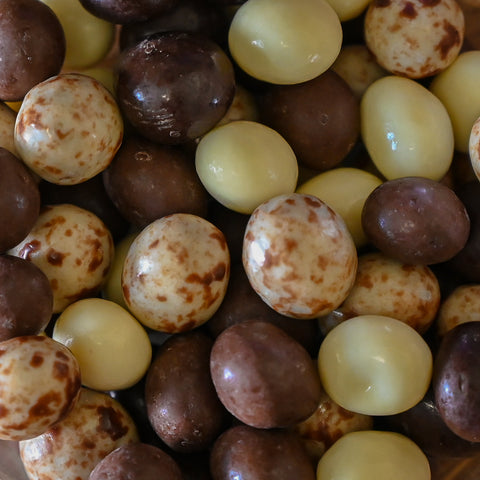 A close-up pile of chocolate-covered round candies, including dark brown chocolate-coated pieces and pale cream chocolate-coated pieces with brown speckles.