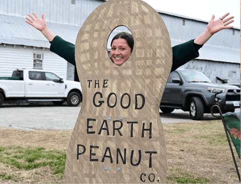 Person peeking out from a cardboard cutout of a peanut shell with 'The Good Earth Peanut Co.' text, outdoors.