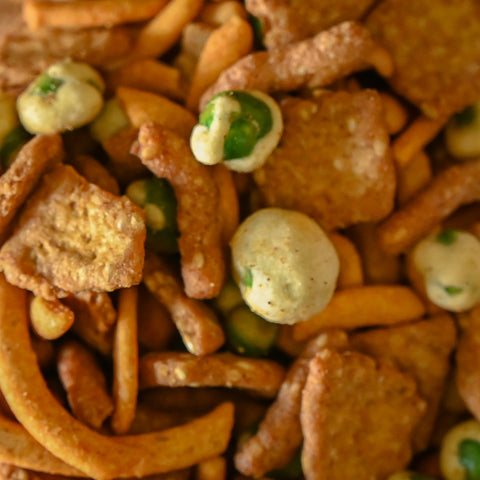 A close-up shot of a snack mix featuring golden-brown pretzel sticks, cracker pieces, peanuts and wasabi covered peas.